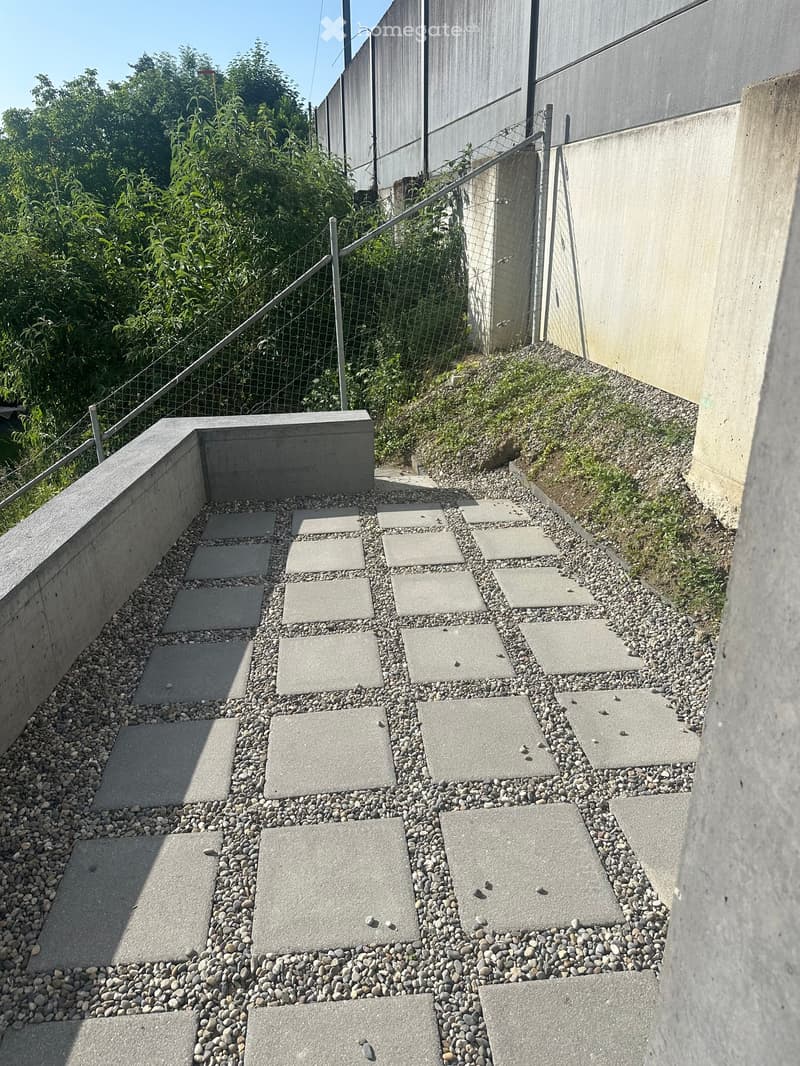 A raised garden area with concrete stepping stones arranged in a pattern, surrounded by small pebbles and bordered by a low concrete wall. A metal railing and greenery are visible in the background.