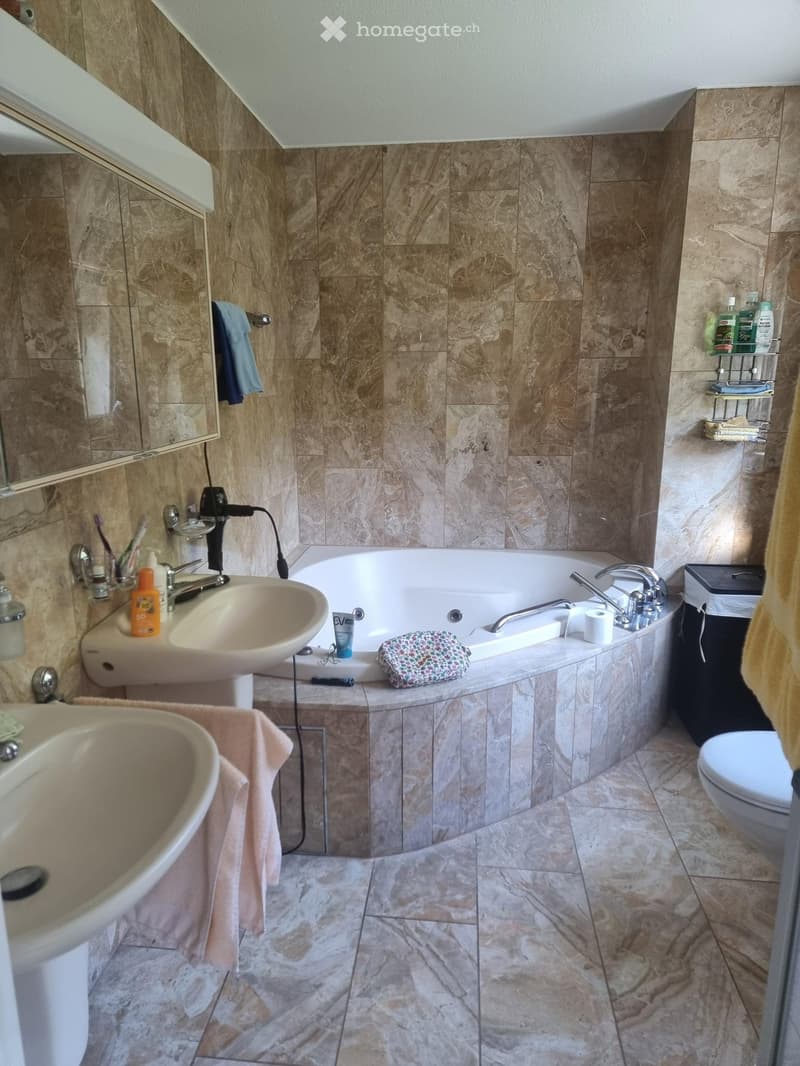 Bathroom with marble tiles, a bathtub, two sinks, a toilet, a mirror, and various toiletries.