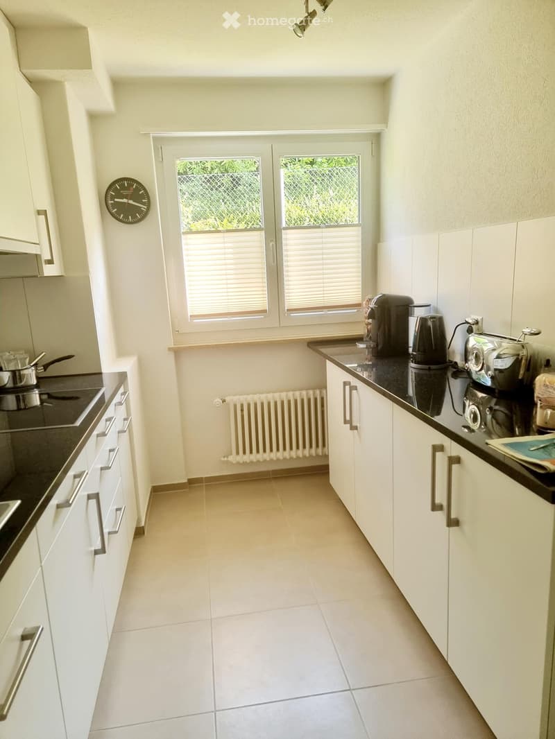 Modern kitchen with white cabinets, black countertops, and stainless steel appliances. Features a coffee maker, toaster, and oven on the counter, along with a clock on the wall and a radiator below the window.