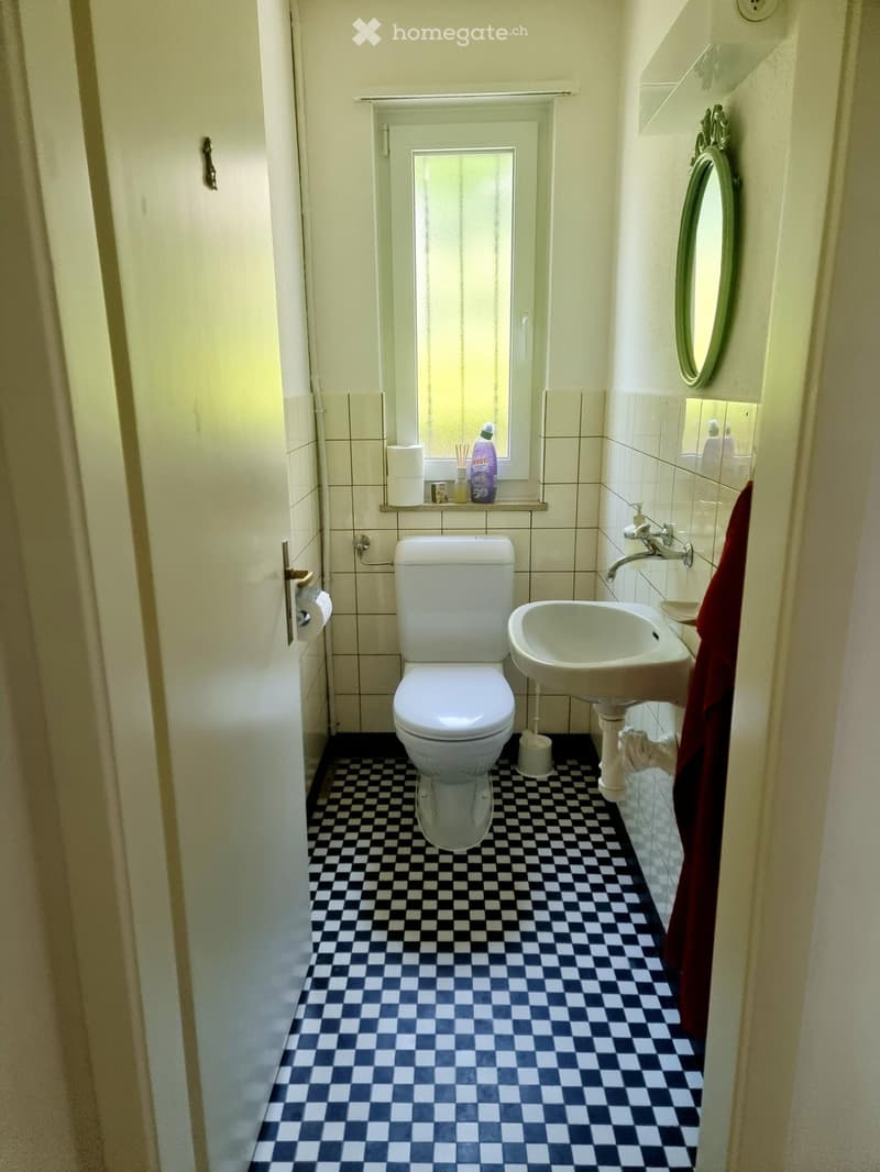 A small bathroom with a black and white checkered floor, a toilet, a sink with a faucet, a mirror, and a towel hanging on the wall. Cleaning supplies are placed on a shelf above the toilet.
