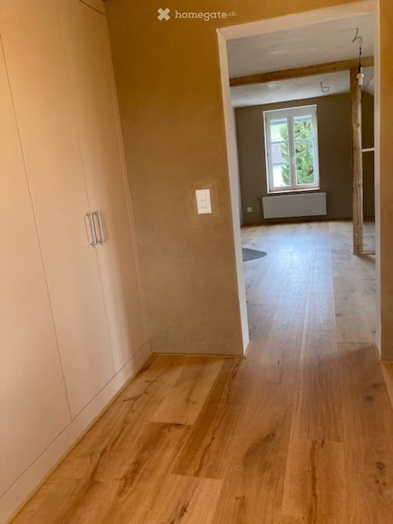 Empty hallway with light wooden flooring, beige walls, and a closed white cabinet on the left. The hallway leads to another room with a window and radiator.