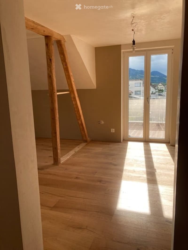Empty room with wooden flooring, exposed wooden beams, large glass door leading to a balcony with a scenic mountain view.