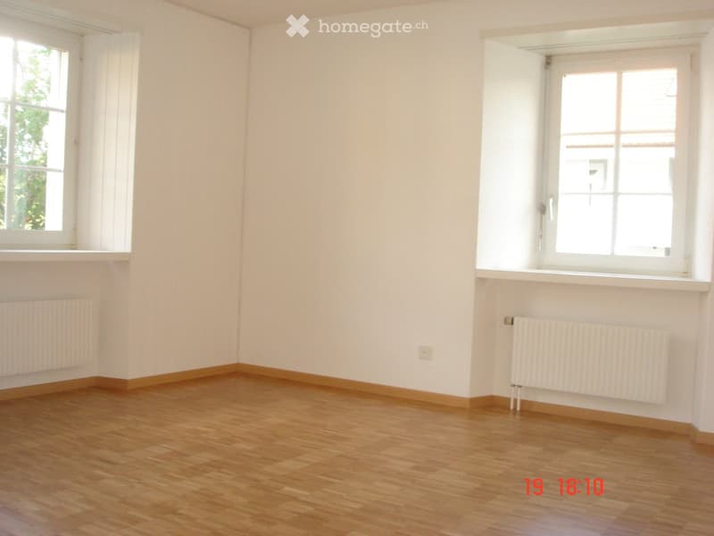 Empty room with wooden flooring, white walls, and two windows with white frames. Radiators are present on both walls.