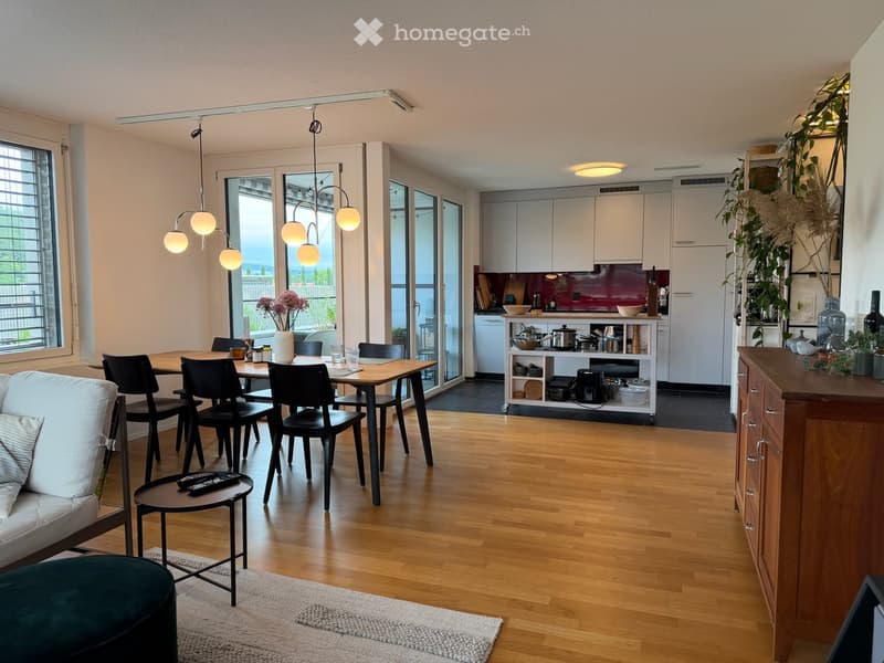 Modern open-plan living and dining area with a sofa, ottoman, coffee table, dining table with chairs, and a fully equipped kitchen with cabinets, appliances, and pendant lighting.