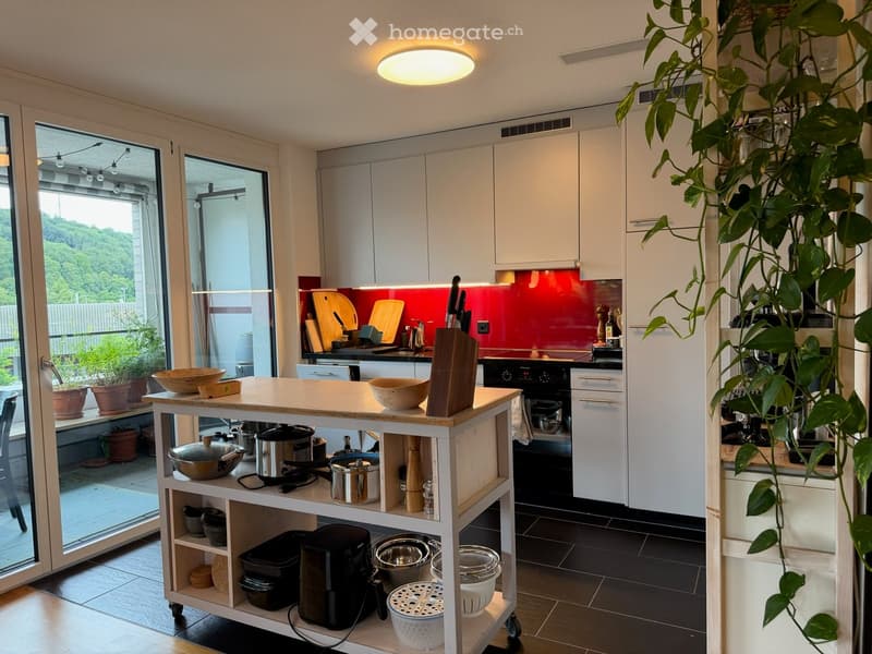 Modern kitchen with white cabinets, a red backsplash, and a central island. The island features bowls, a cutting board, and various kitchen utensils. A large plant is visible on the right side, and the kitchen has large windows with a view of greenery out