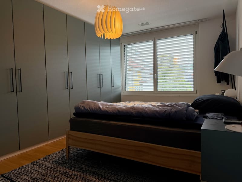 Modern bedroom with a wooden bed, nightstand, wardrobe, and a hanging lamp. The room features wooden flooring and large windows with blinds.