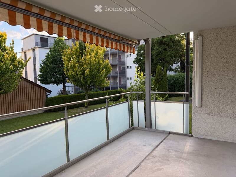 Balcony with a striped awning, metal railing, and a view of a residential area with trees and buildings.