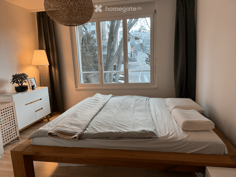large window with a city view, wooden bed with white sheets, potted plant, side table with lamp, white dresser