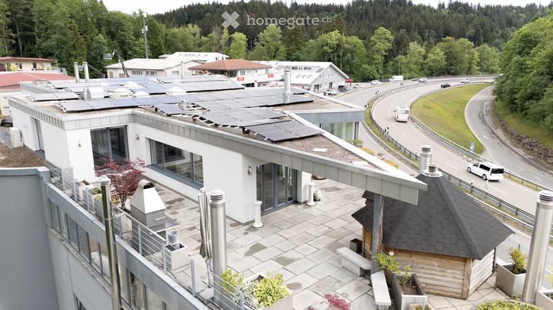 Modern building with solar panels on the roof, surrounded by greenery and a road nearby.