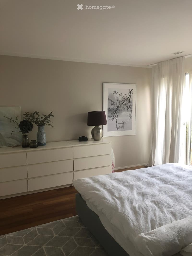 A modern bedroom with a large bed, nightstand, lamp, vase with flowers, picture frame, and mirror on the wall.
