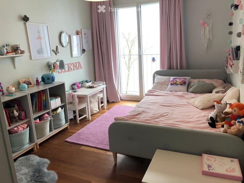 A well-lit bedroom with a bed, a small table, a shelf, and various decorative items including a dreamcatcher and framed pictures. The room features a pink rug and a window with curtains.