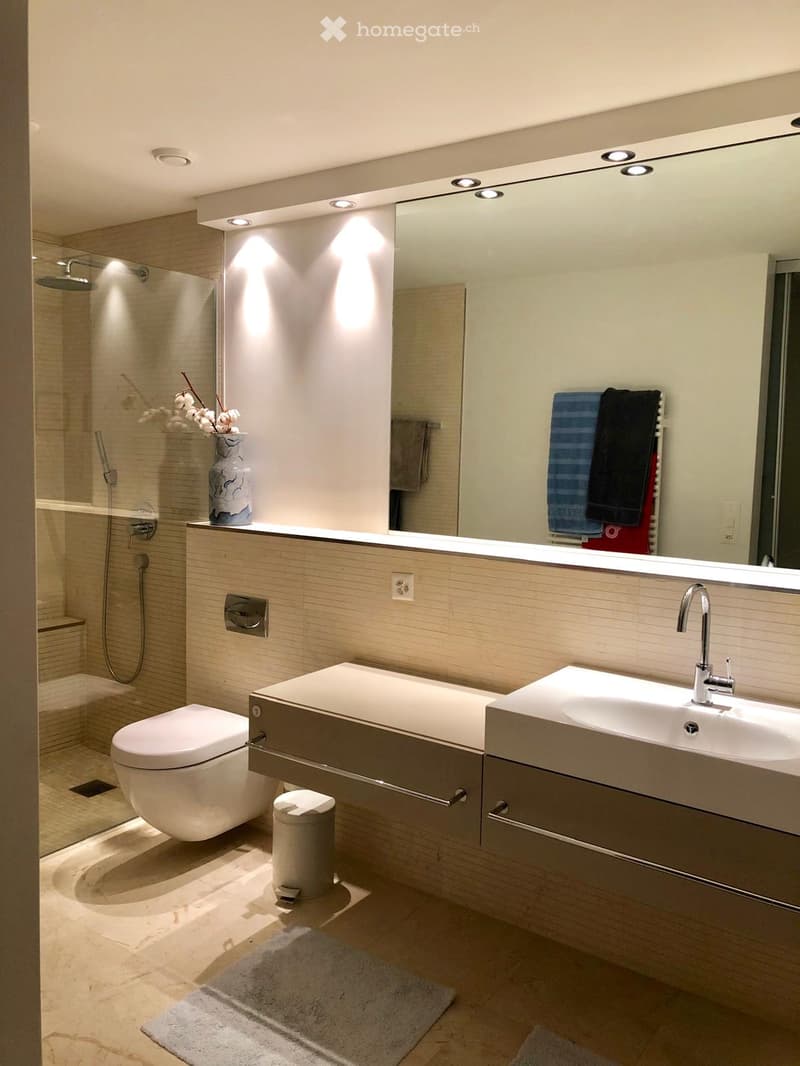 Modern bathroom with a large mirror, double sink, toilet, and a glass-enclosed shower area. Features include built-in lighting, a sleek vanity, and decorative towels.