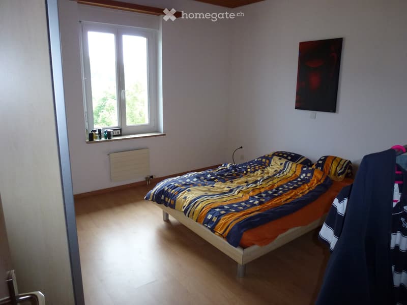 Bedroom with wooden floor, bed with blue and yellow bedding, radiator, painting on wall, and wardrobe with clothes.