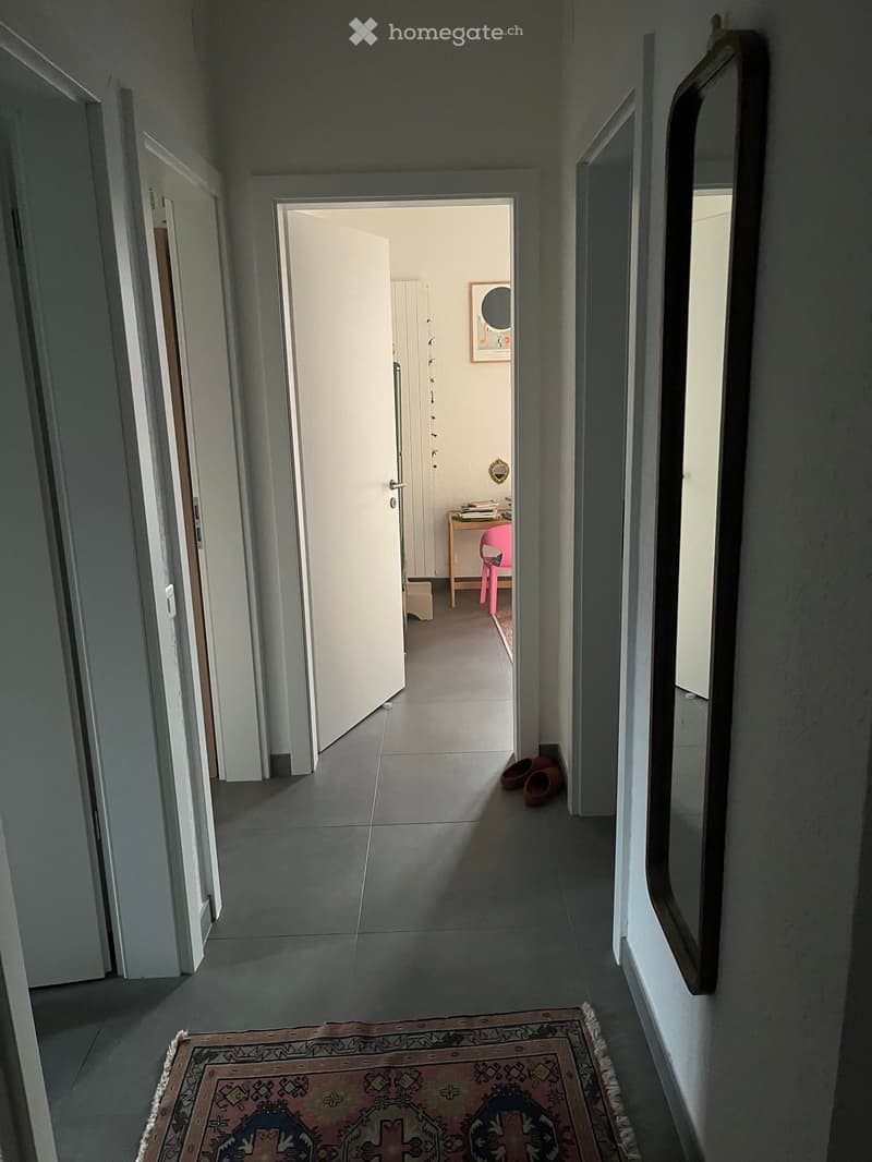 A long, narrow hallway with grey tiled flooring, white walls, and several open doors leading to other rooms. A small pink chair and a pair of brown shoes are visible in the adjacent room.