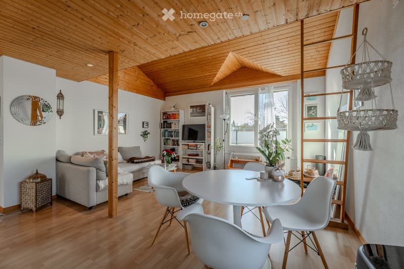 A modern living room with wooden floors, exposed wooden beams, a grey sofa, a white dining table with chairs, a bookshelf, and a television. The room features a hanging decorative chandelier and a large window with a view of the outdoors.