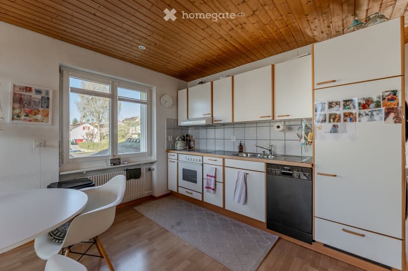 Bright kitchen with white cabinets, wooden ceiling, and large windows. Includes a refrigerator, oven, dishwasher, sink, and a small dining area with a table and chairs.