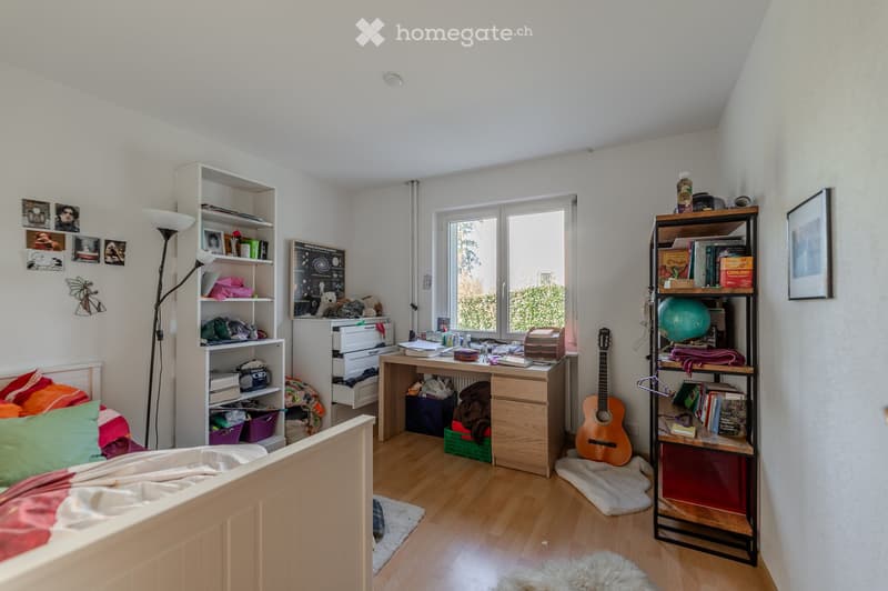 A cozy bedroom with a wooden bed, colorful pillows, a desk with various items, shelves filled with books and decorative objects, a guitar, and a globe. The room features wooden flooring and large windows letting in natural light.