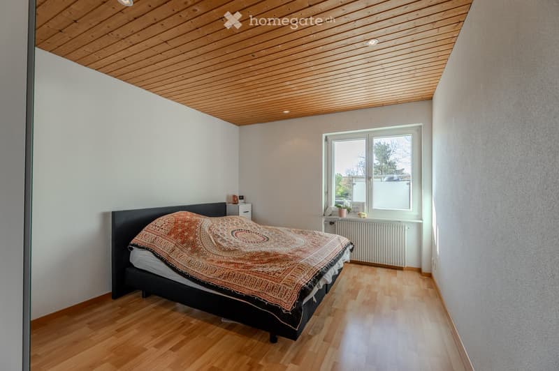 Bright bedroom with wooden flooring, a large window, a bed with a patterned bedspread, a nightstand, and a radiator beneath the window.