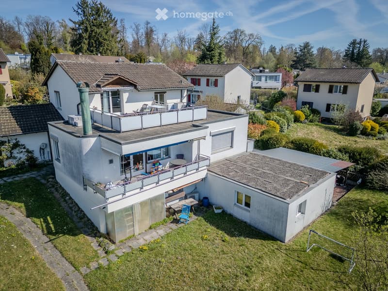 Aerial view of a modern two-story house with a flat roof, surrounded by a well-maintained garden and neighboring houses. Features include a balcony, patio area, and outdoor furniture.