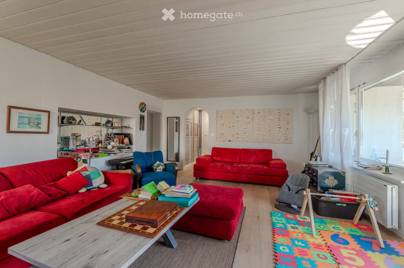 Bright living room with red sofas, a coffee table, ottoman, armchair, baby chair, and colorful play mat. Shelves and artwork are visible on the walls.