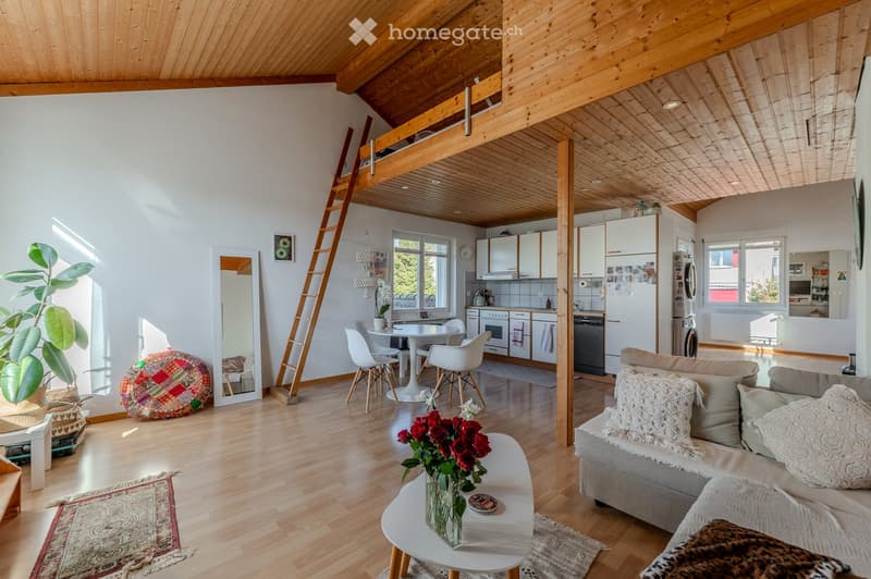 Bright, open-concept living and dining area with wooden floors, white walls, and a slanted wooden ceiling. Features a grey sofa with decorative pillows, a white coffee table with a flower vase, a dining table with white chairs, a ladder leading to a loft