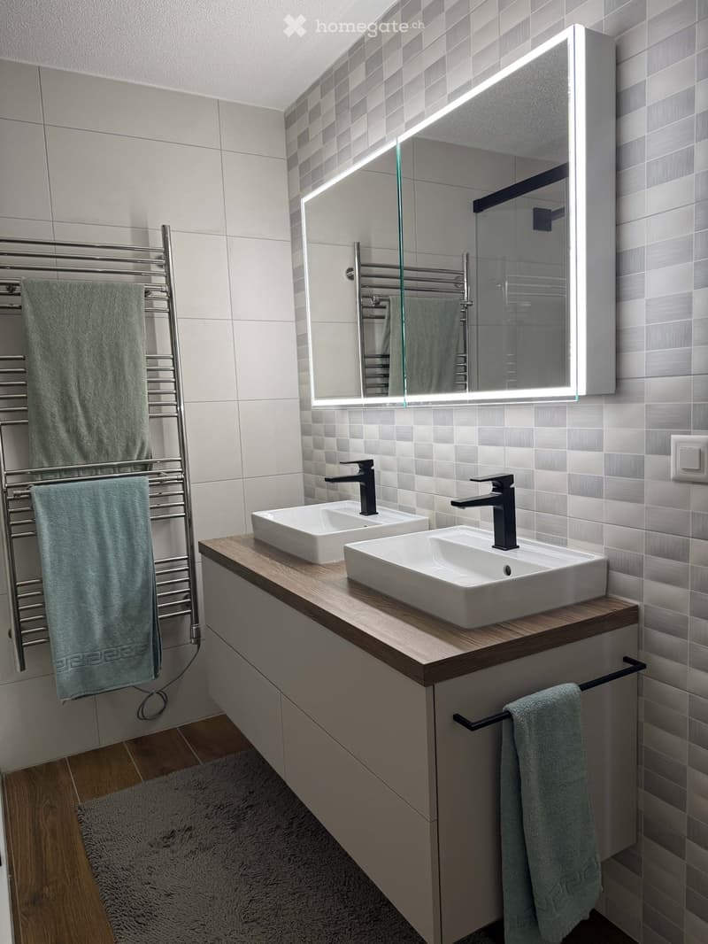 Modern bathroom with two white sinks, sleek black faucets, a large illuminated mirror, a towel rack with green towels, and a wooden countertop.