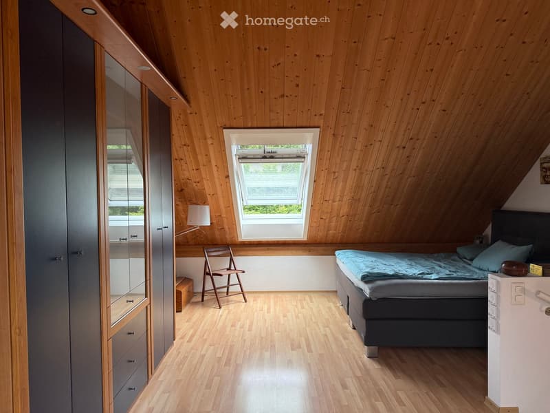 Cozy attic bedroom with wooden walls and ceiling, featuring a bed, a chair, a wardrobe, and a lamp.