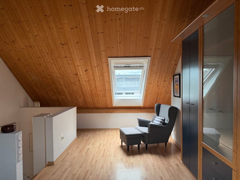 Attic room with wooden walls and ceiling, skylight window, grey armchair, ottoman, and modern cabinet.