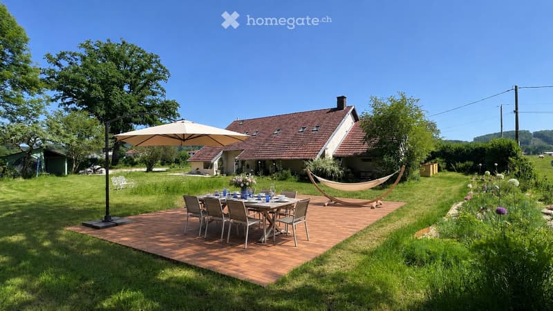 A wooden deck with a dining table, chairs, and an umbrella, surrounded by a grassy lawn with a hammock and a bench, with a house in the background.