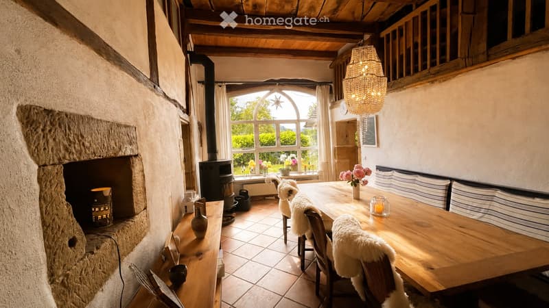 Cozy dining area with a large wooden table, chairs with fur cushions, a stone fireplace, and a chandelier hanging from the wooden ceiling.