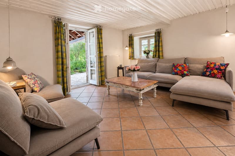 Cozy living room with a large sectional sofa, coffee table, and a view to the garden through an open door. The room features tiled flooring, hanging pendant lights, and decorative pillows on the sofa.