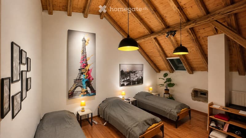 Cozy attic bedroom with wooden beams, three single beds, nightstands, table lamps, artwork, and a potted plant.