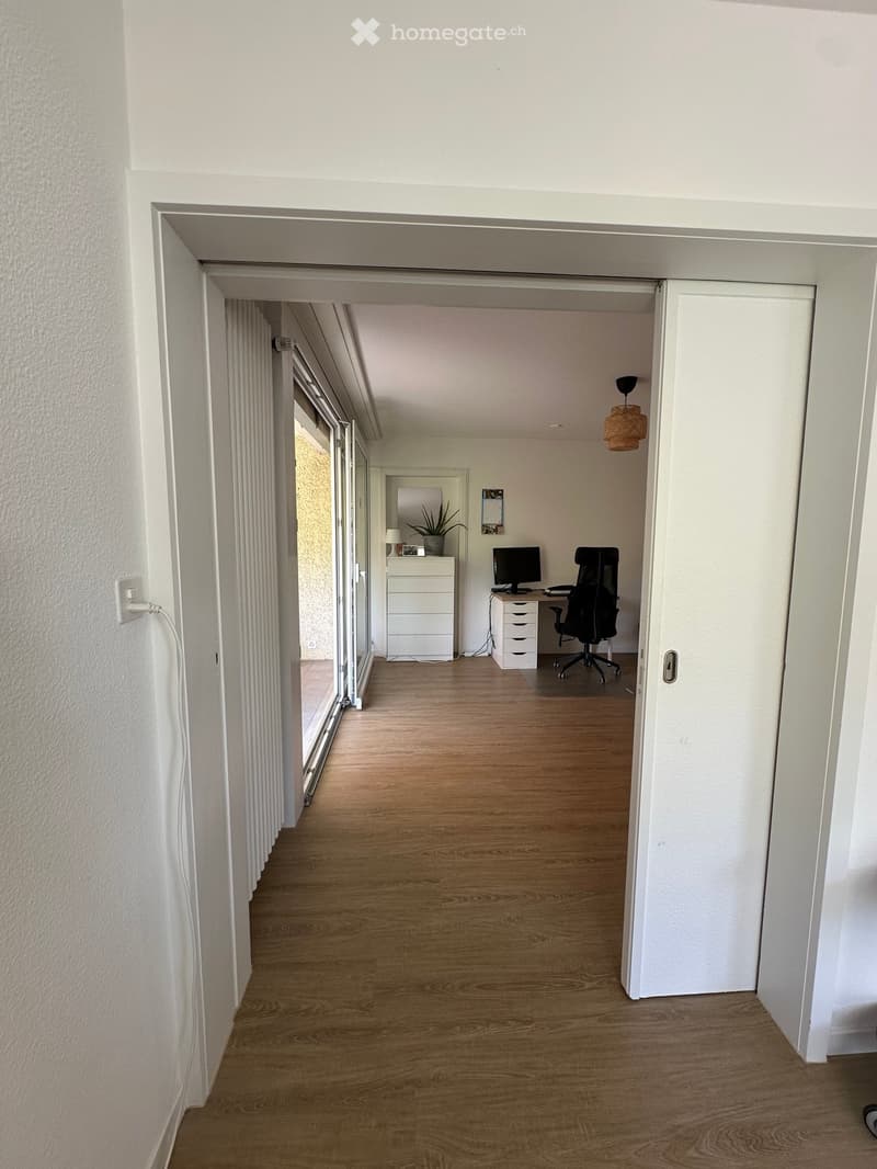 A clean, modern hallway with wooden flooring, white walls, and a hanging woven pendant light. An open doorway leads to a furnished room with a desk, chair, and computer.