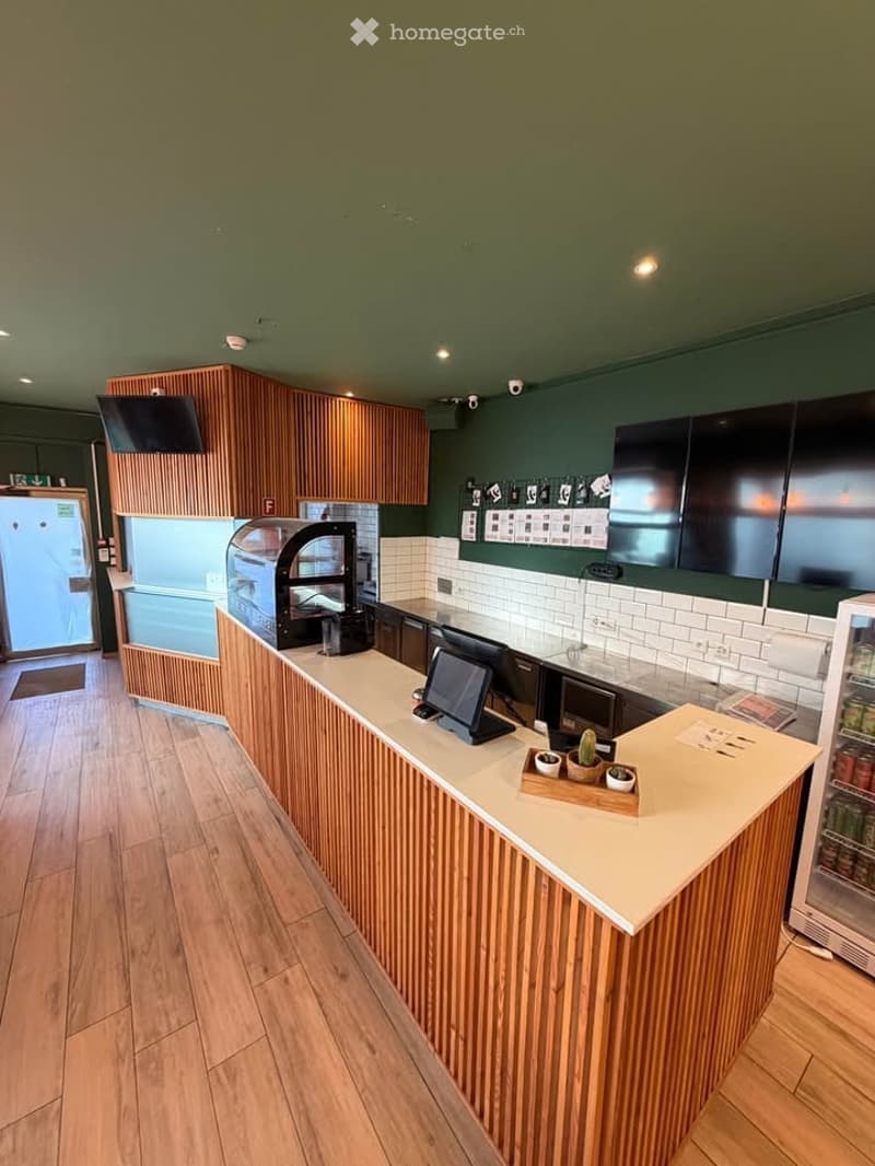 Modern kitchen area with wooden cabinets, a central counter, and a display case. Includes screens, a refrigerator, and small decorative items on the counter.