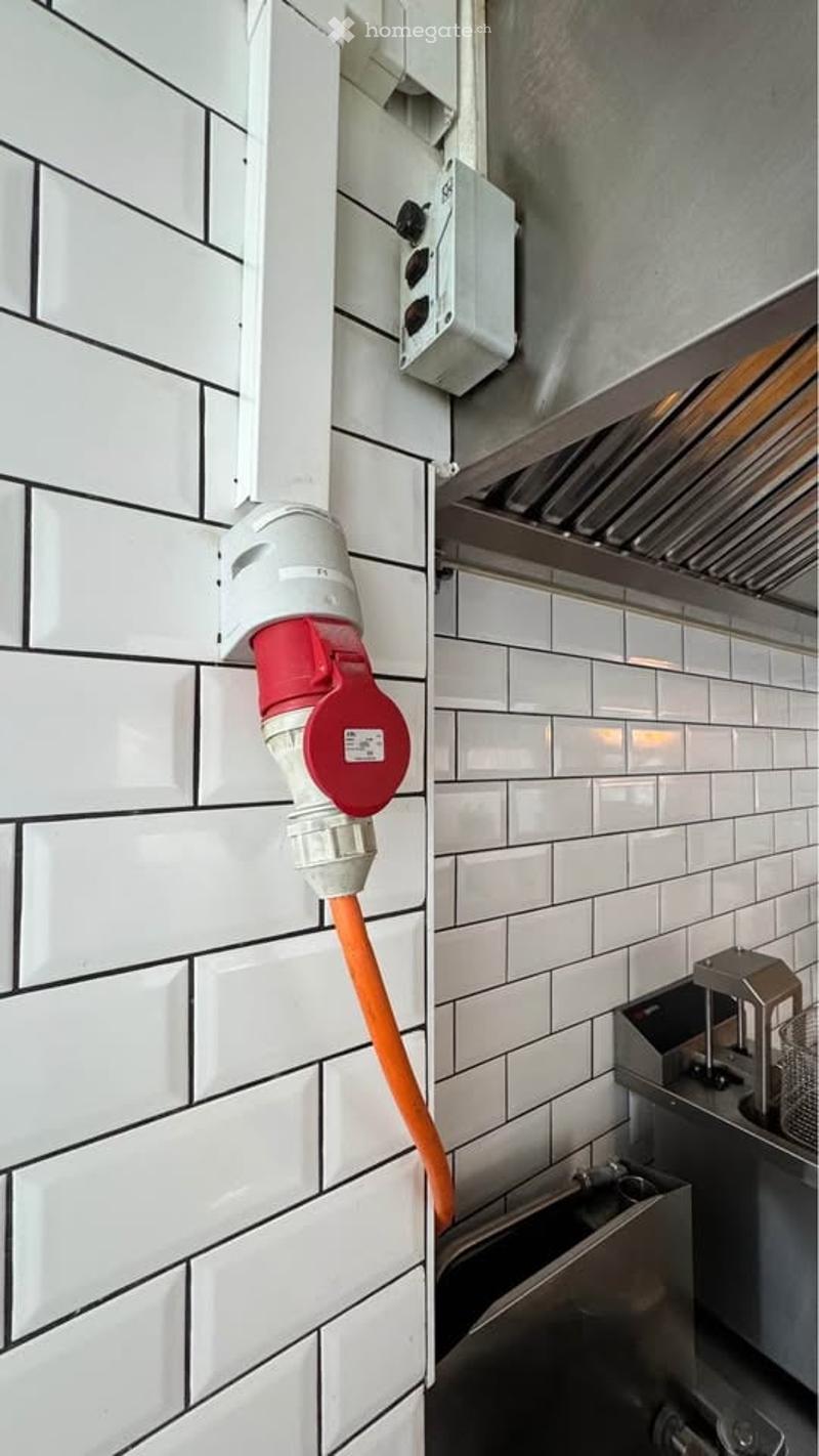 A close-up view of a kitchen wall with a white tiled surface, featuring a red and white electrical outlet and a grey kitchen hood in the background.