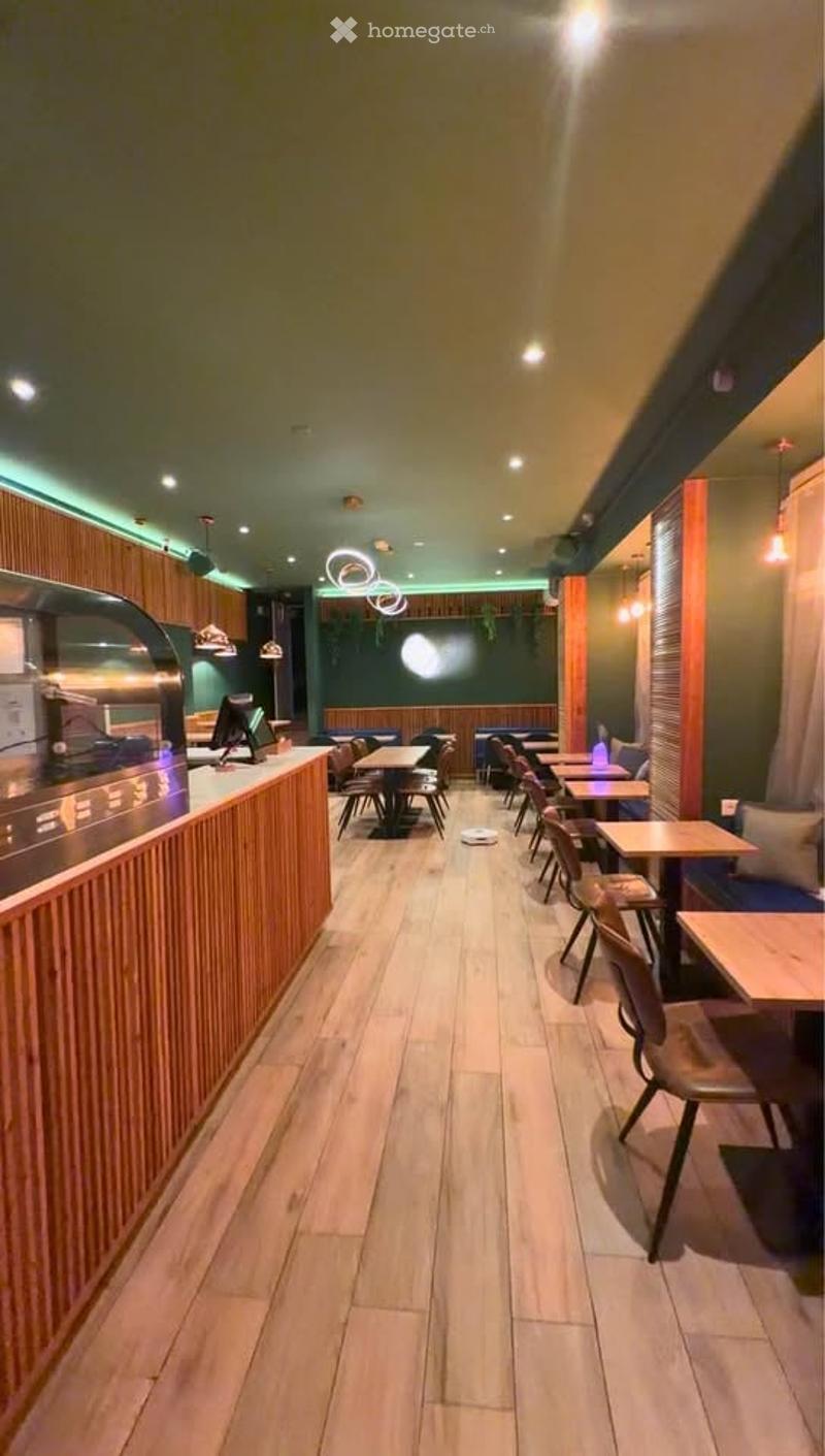 Modern restaurant interior with wooden flooring, a counter with a screen, and multiple tables and chairs arranged in rows.