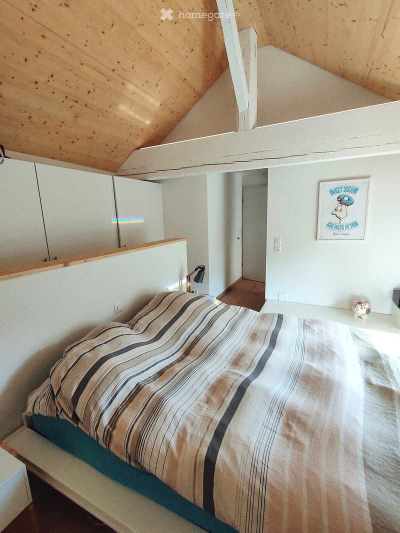 Cozy attic bedroom with a slanted wooden ceiling, a single bed with striped bedding, a wall-mounted lamp, a mirror, and a poster on the wall.