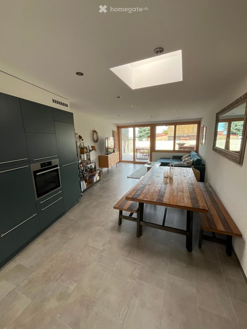 Open-plan kitchen and dining area with a large wooden dining table, benches, a sofa, dark green cabinets, and a modern oven. Large windows and a skylight provide natural light.