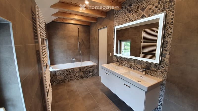 Modern bathroom with a large mirror, double sink, bathtub, and radiator. The walls are tiled with a decorative pattern, and the floor is tiled in a warm brown tone.