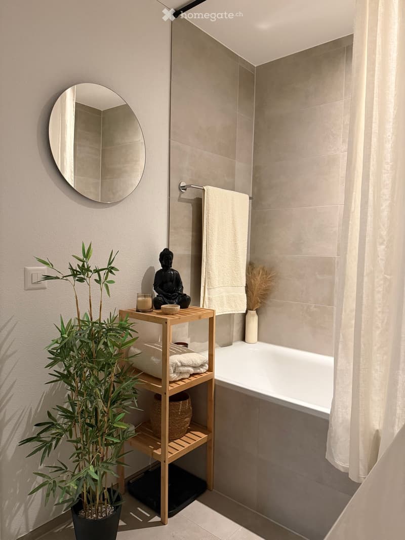 Modern bathroom with a bathtub, wooden shelf holding towels, a potted plant, a decorative statue, and a round mirror on the wall.