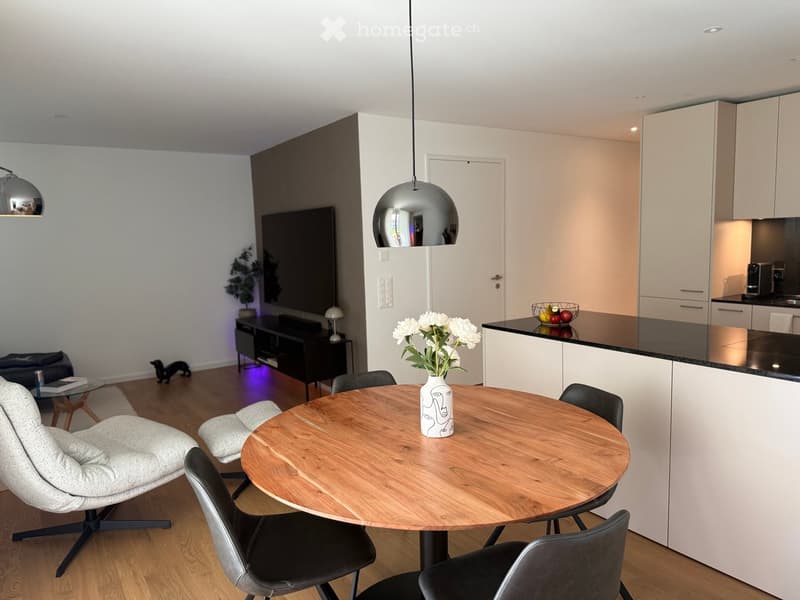 Modern living and dining area with a round wooden table, chairs, sofa, TV, kitchen area with cabinets and counter, and decorative items including a vase with flowers and a stool.