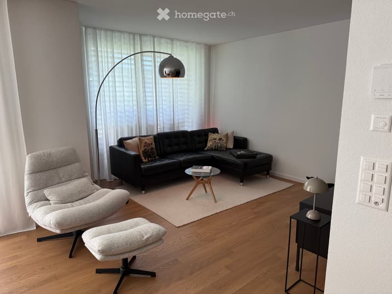 Modern living room with a black sectional sofa, a grey armchair, a matching ottoman, a glass coffee table, and a floor lamp. The room features wooden flooring and large windows with white curtains.