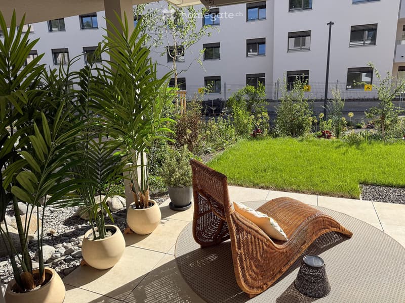 A modern outdoor patio with a wicker lounge chair, small table, and several potted plants, surrounded by a well-maintained lawn and modern building.
