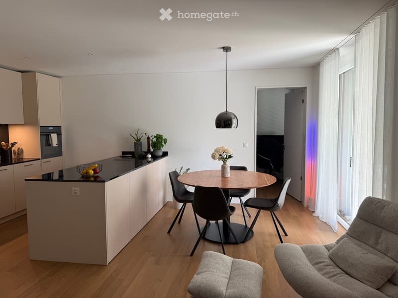 Modern open-plan living and dining area with a grey sofa, round wooden dining table with four chairs, kitchen island with built-in oven, pendant light, and decorative vases.