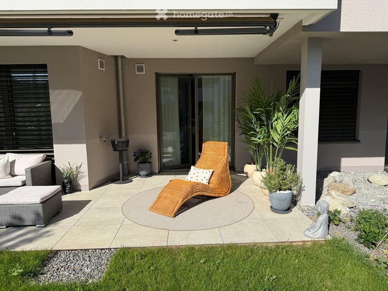 A modern outdoor patio with a grey wicker sofa, a lounge chair, a small table, and several potted plants, including tall leafy plants and smaller shrubs. The area is tiled and features a small garden with grass and rocks.