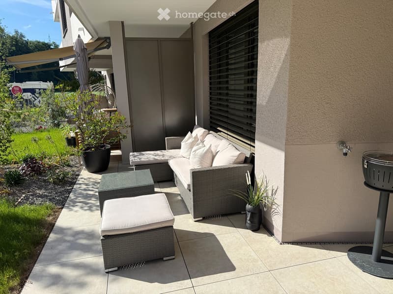 A modern outdoor patio with a grey wicker sofa, matching stool, small table, umbrella, and barbecue, surrounded by potted plants and a well-maintained garden.