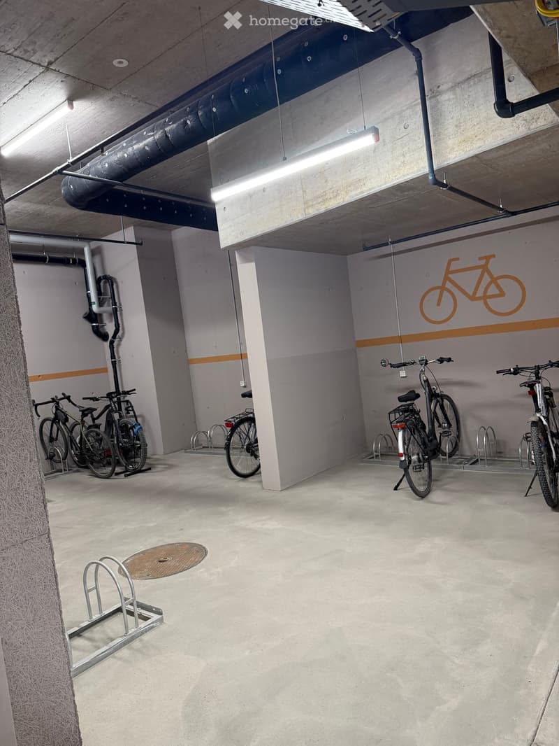 Underground parking area with several bicycles parked on racks, concrete floor, exposed pipes, and lighting.