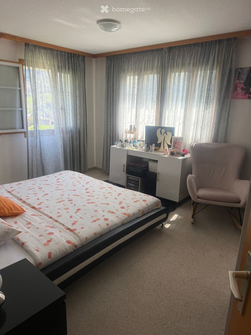 A well-lit bedroom with a modern bed, a pink armchair, a desk with a screen, and a cabinet. The room features large windows with curtains and a carpeted floor.