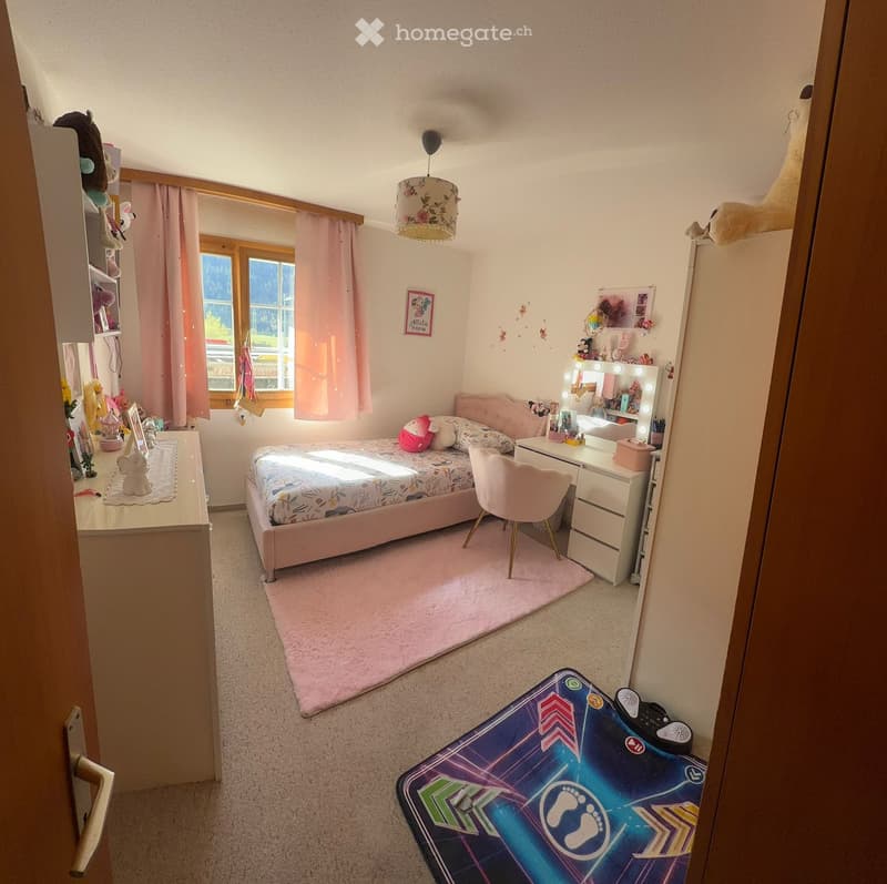A well-lit bedroom with a pink bed, a vanity desk with a mirror, a pink chair, and a colorful play mat on the floor. The room features a large window with pink curtains and a shelf with toys and decorations.