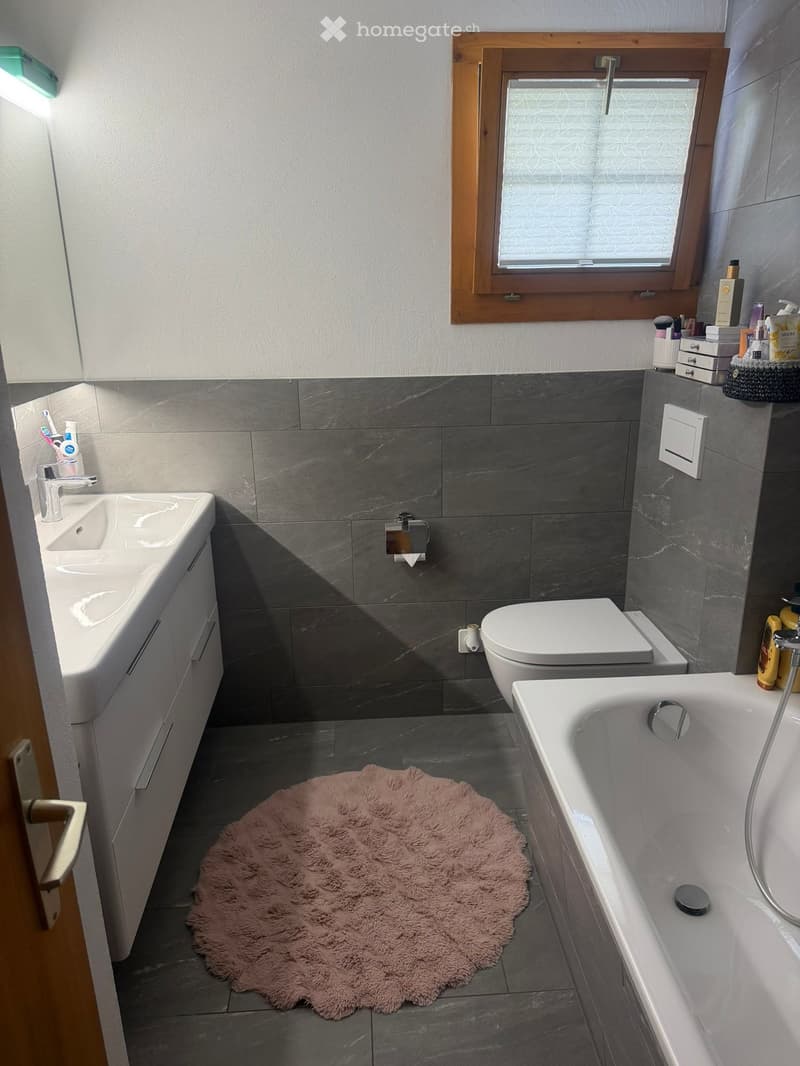 Modern bathroom with grey tiles, white fixtures, a bathtub, toilet, sink, and a pink bath mat on the floor.